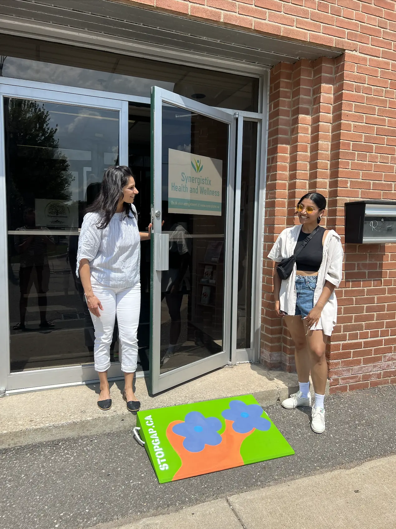 Saphera and the business owner beside a painted ramp with blue flowers on green and orange, at a brick storefront entrance in downtown Brampton