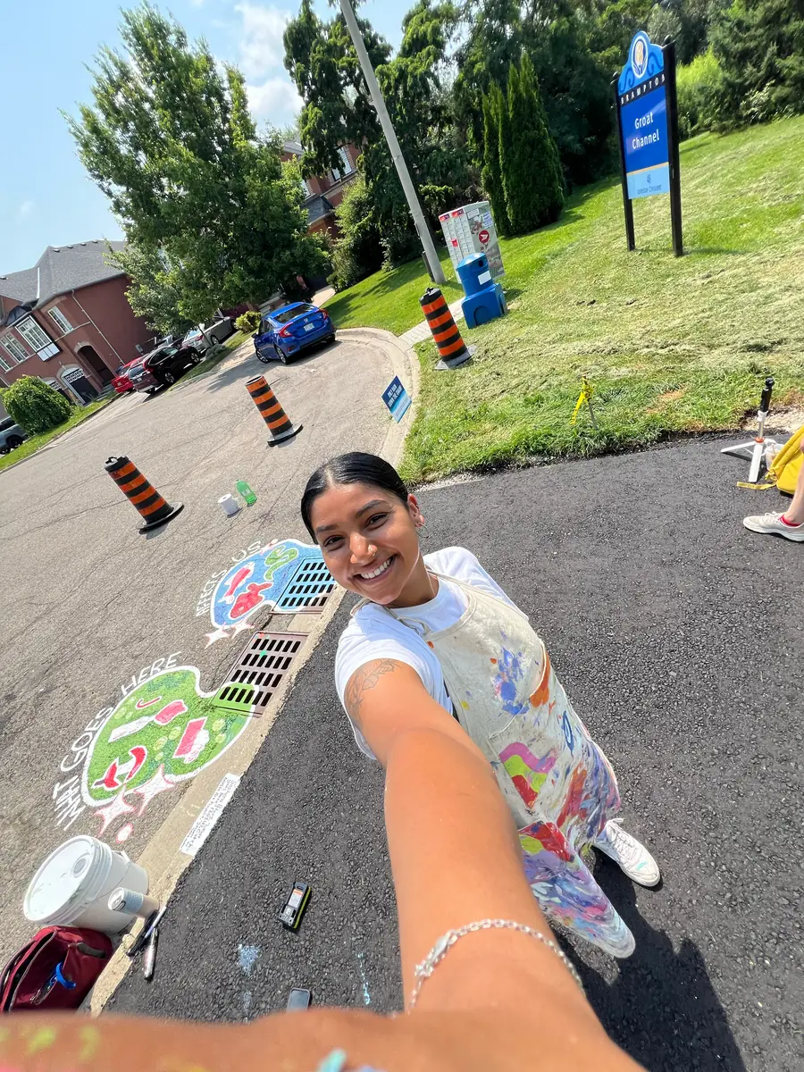 Saphera reaching toward the camera beside the second catch basin mural on a sunny street
