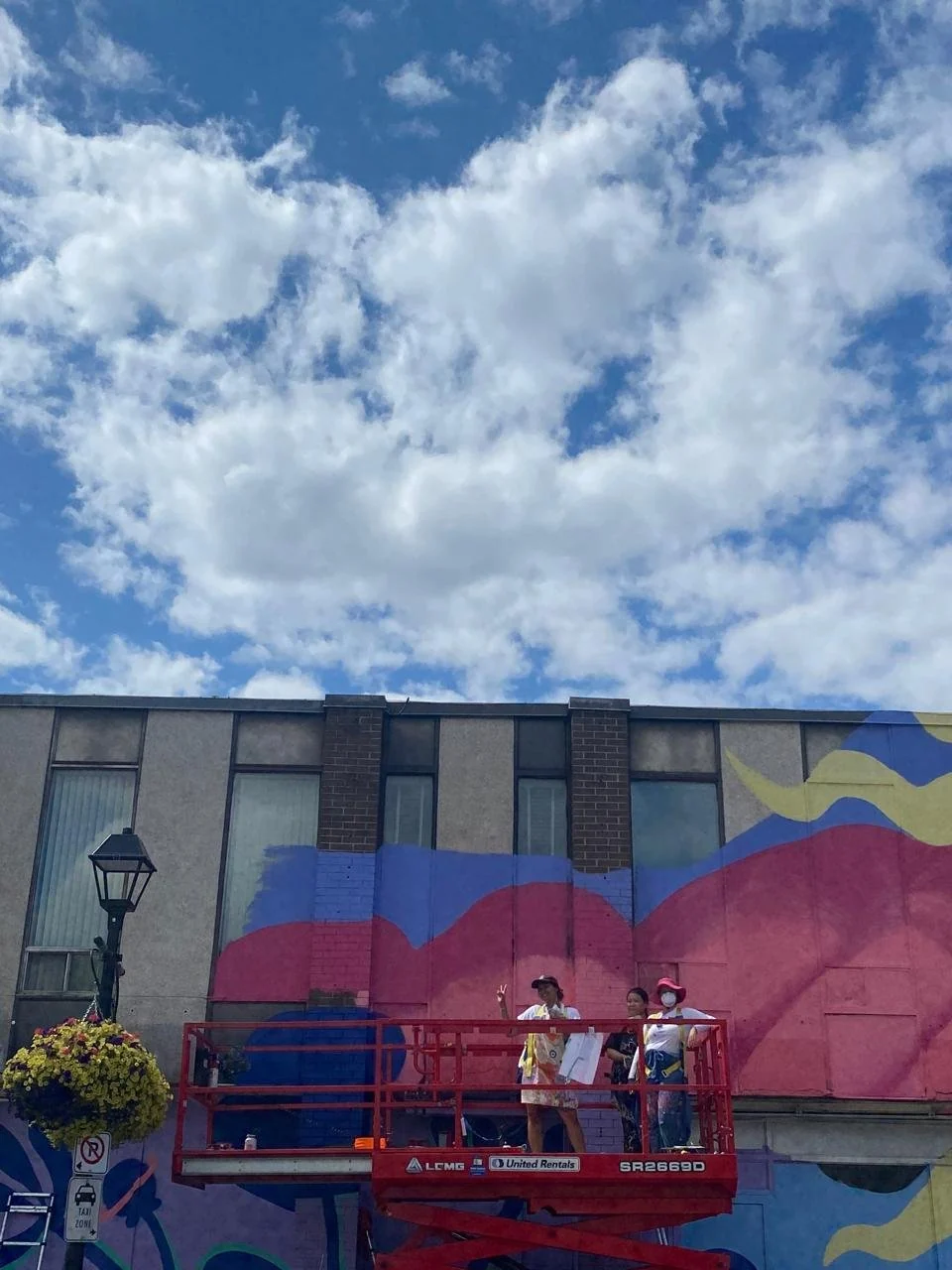 Wide view of the scissor lift against the mural under a bright cloudy sky