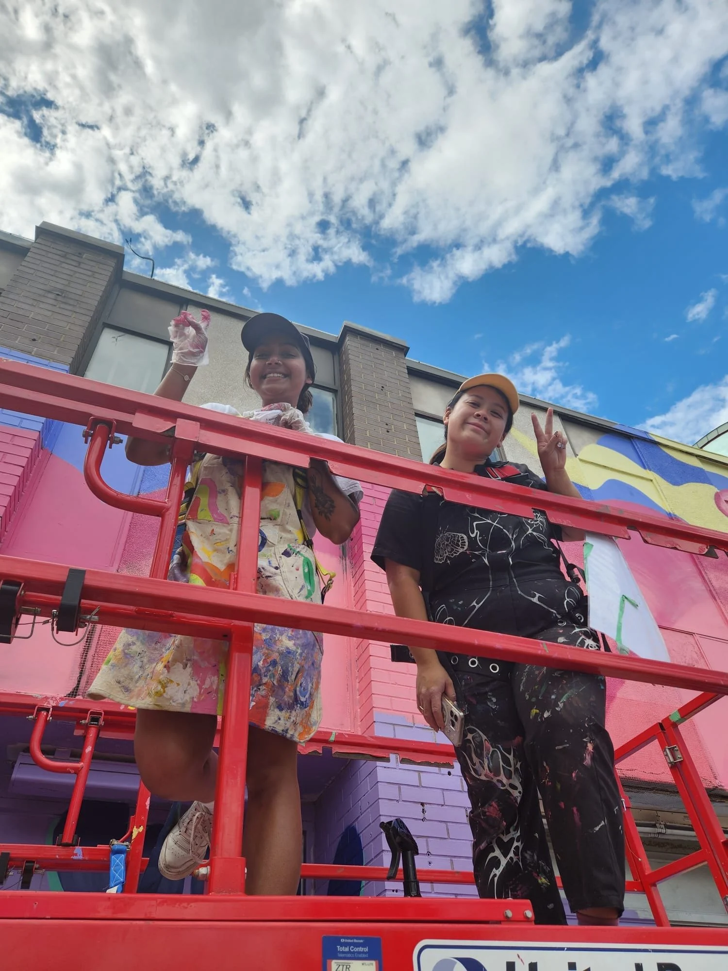 Two artists on a red scissor lift painting the upper section of the mural