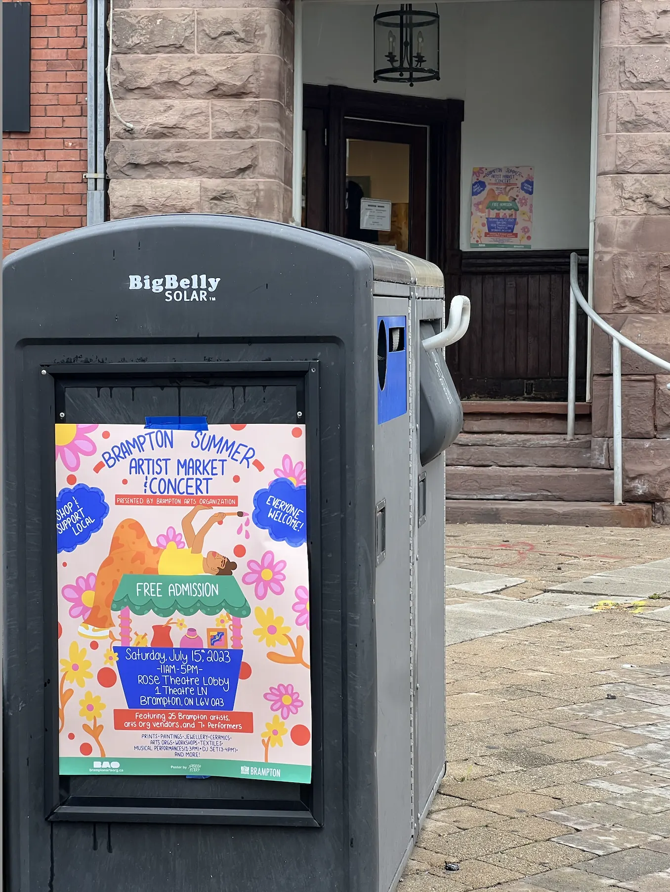 The Summer Artist Market poster displayed on a BigBelly bin outside a stone building