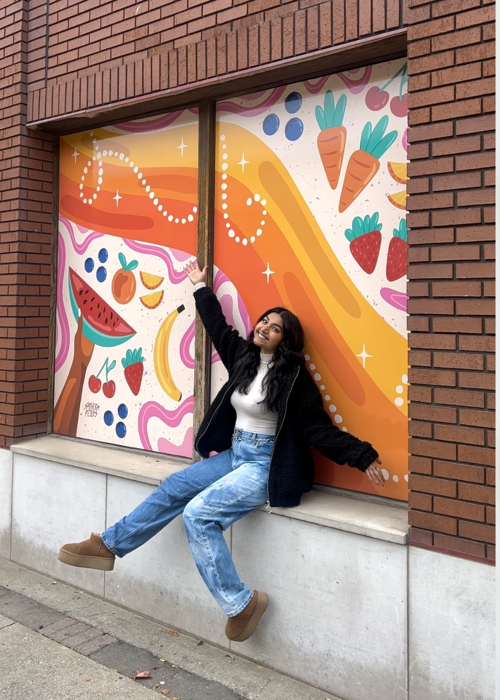 Saphera leaning against a storefront window wrapped in an orange and pink arts and culture mural